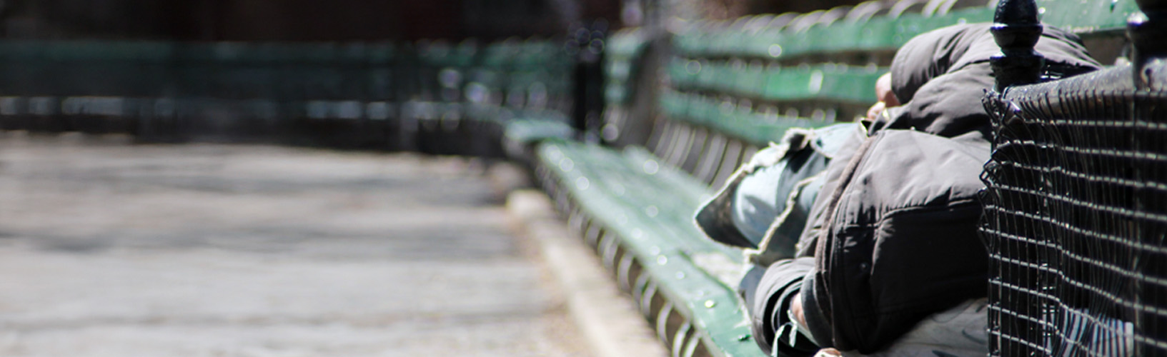 Homeless person in hooded jacket sleeping on park bench in New York City. 